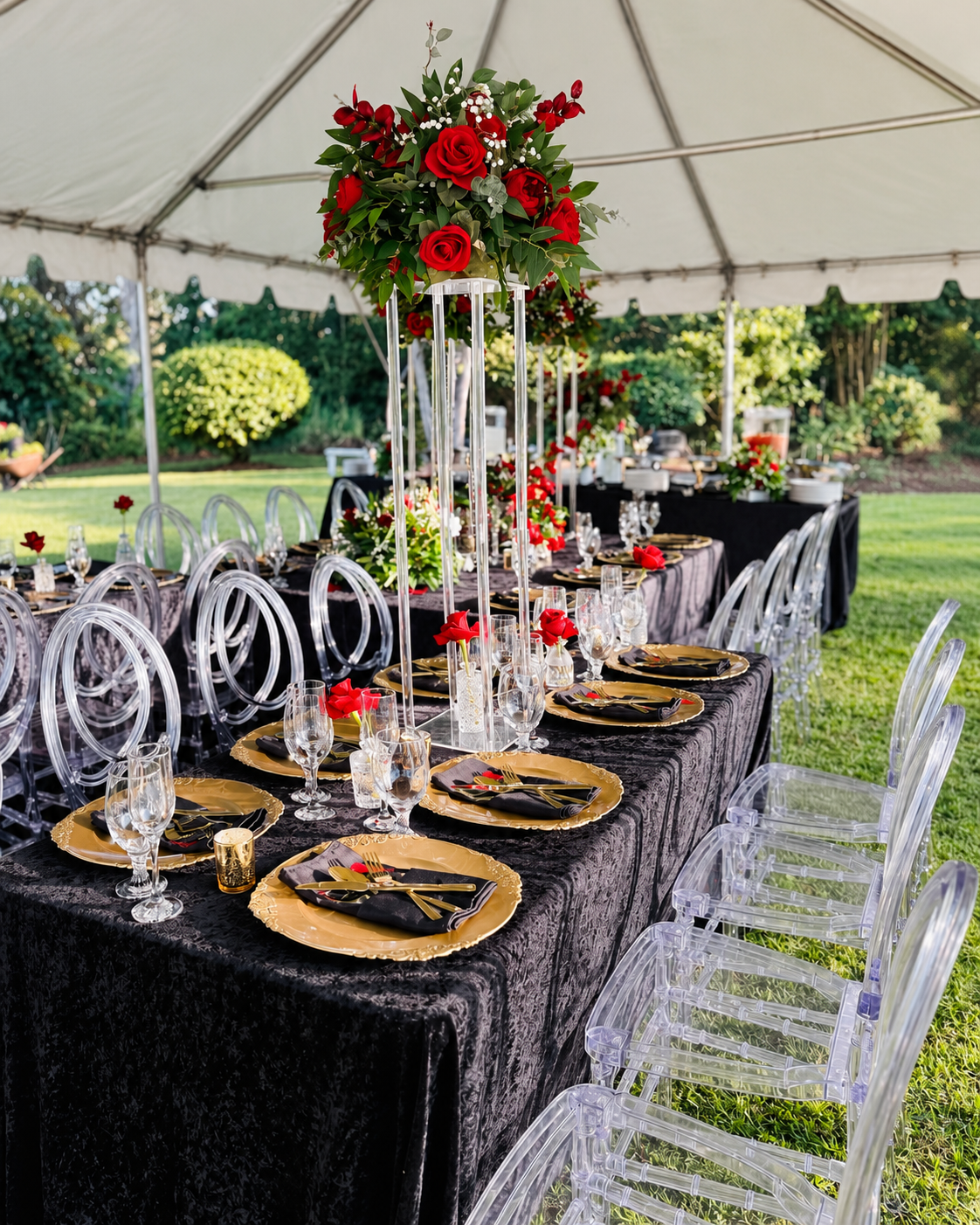 Garden reception table with red rose centerpieces, gold place settings, and clear chairs.